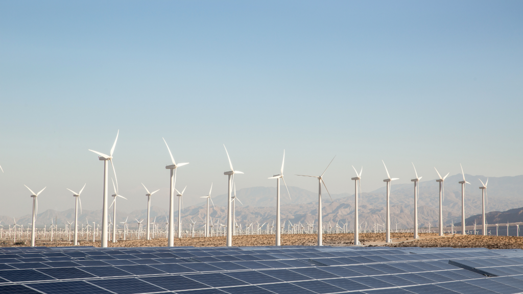 Rows of white wind turbines stand behind solar panels in a desert landscape, symbolizing sustainable wealth in harmony with nature, with mountains and a clear blue sky in the background.