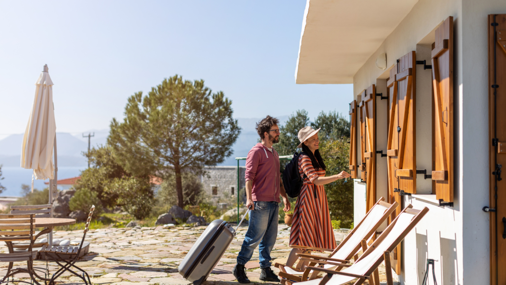 A man and woman arrive at a vacation rental with suitcases. The woman unlocks the door while the man stands beside her, enjoying the sense of wealth and relaxation. Deck chairs and an outdoor table sit on a patio, framed by trees and mountains.