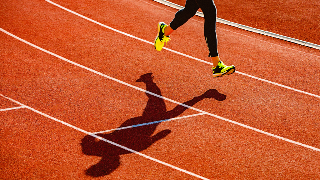 A runner wearing black leggings and bright yellow shoes is captured mid-stride on a red running track, embodying the focus and determination needed for successful financial planning. Their shadow is clearly cast on the surface.
