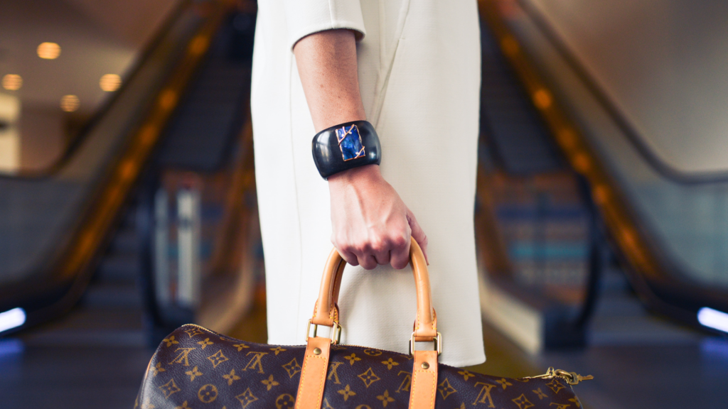 A person in a white outfit stands in front of escalators, holding a brown Louis Vuitton bag and wearing a large black smartwatch, exuding wealth in an upscale indoor setting.
